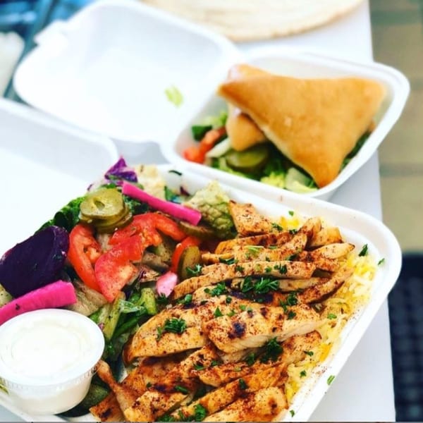 Two takeout containers of Mediterranean food, with grilled chicken, rice, salad, and a pastry.
