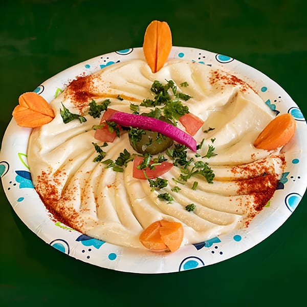 Creamy hummus garnished with parsley, tomatoes, pickles, paprika, and carrot slices on a paper plate.