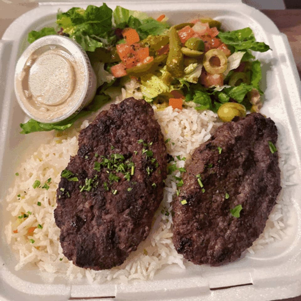 Two grilled Mediterranean beef patties with rice, salad, and a side of sauce in a takeout container.